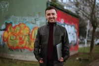 Филип Наков со својот лаптоп стои насмеан пред графит